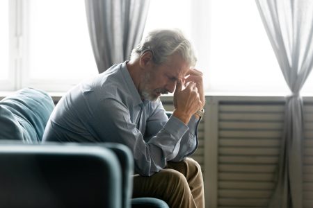 Side view in living room seated on couch hunched old 50s male hold hands near face looking desperate and lonely feels worried. Concept of illness and diagnosis, older generation humans life troubles