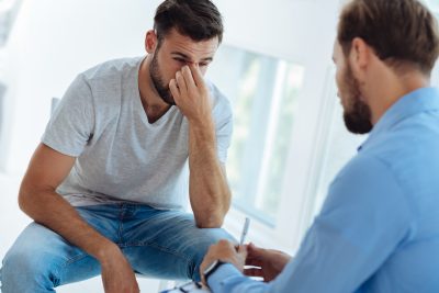 Sadness and depression. Unhappy nice young man holding the bridge of his note and thinking about his problems while having a talk with a psychologist
